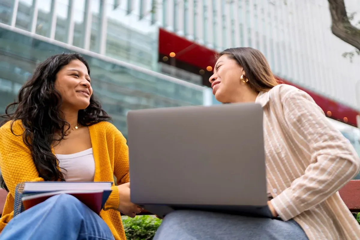 Two college students sit outside with a laptop and books, smiling and talking on campus.